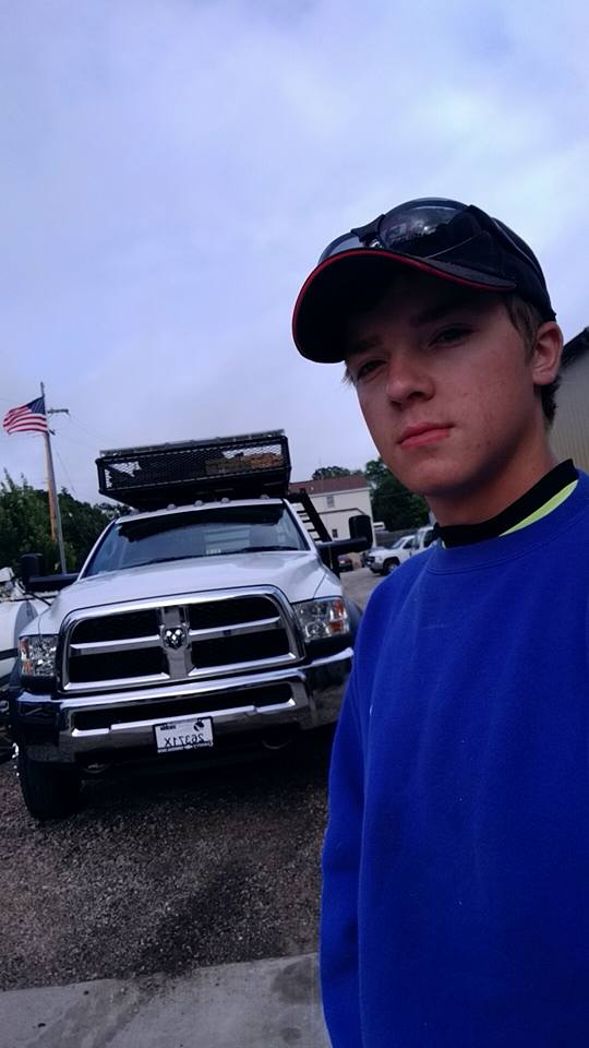 Person wearing a blue sweatshirt and cap standing in front of a white truck with a toolbox on a cloudy day.