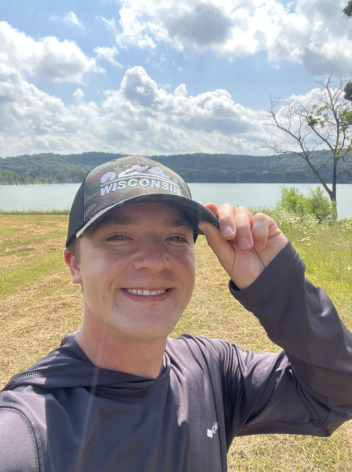 Man wearing a 'Wisconsin' cap in a scenic outdoor setting with a lake and mountains.