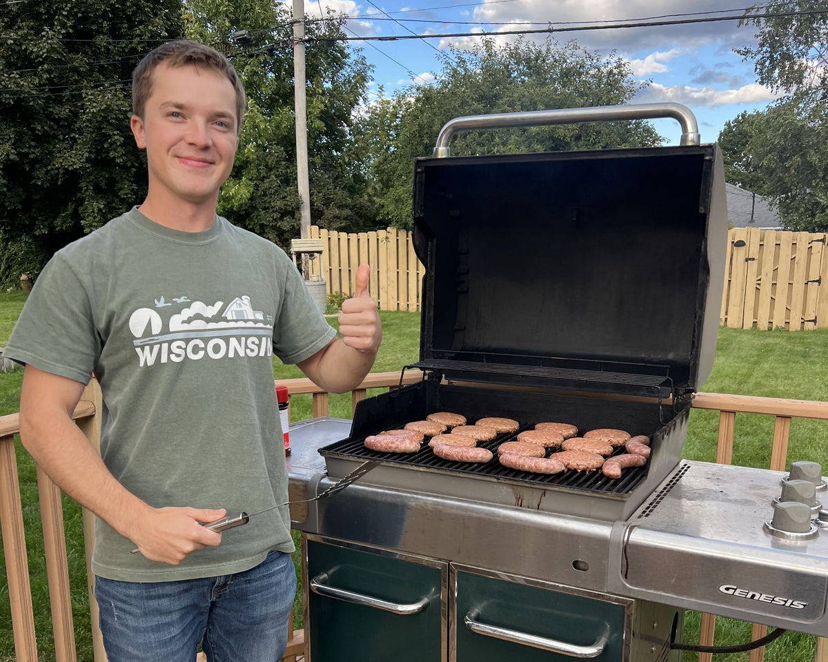 Man grilling outdoors on a deck with a grill and propane tank while wearing a "Wisconsin" shirt.