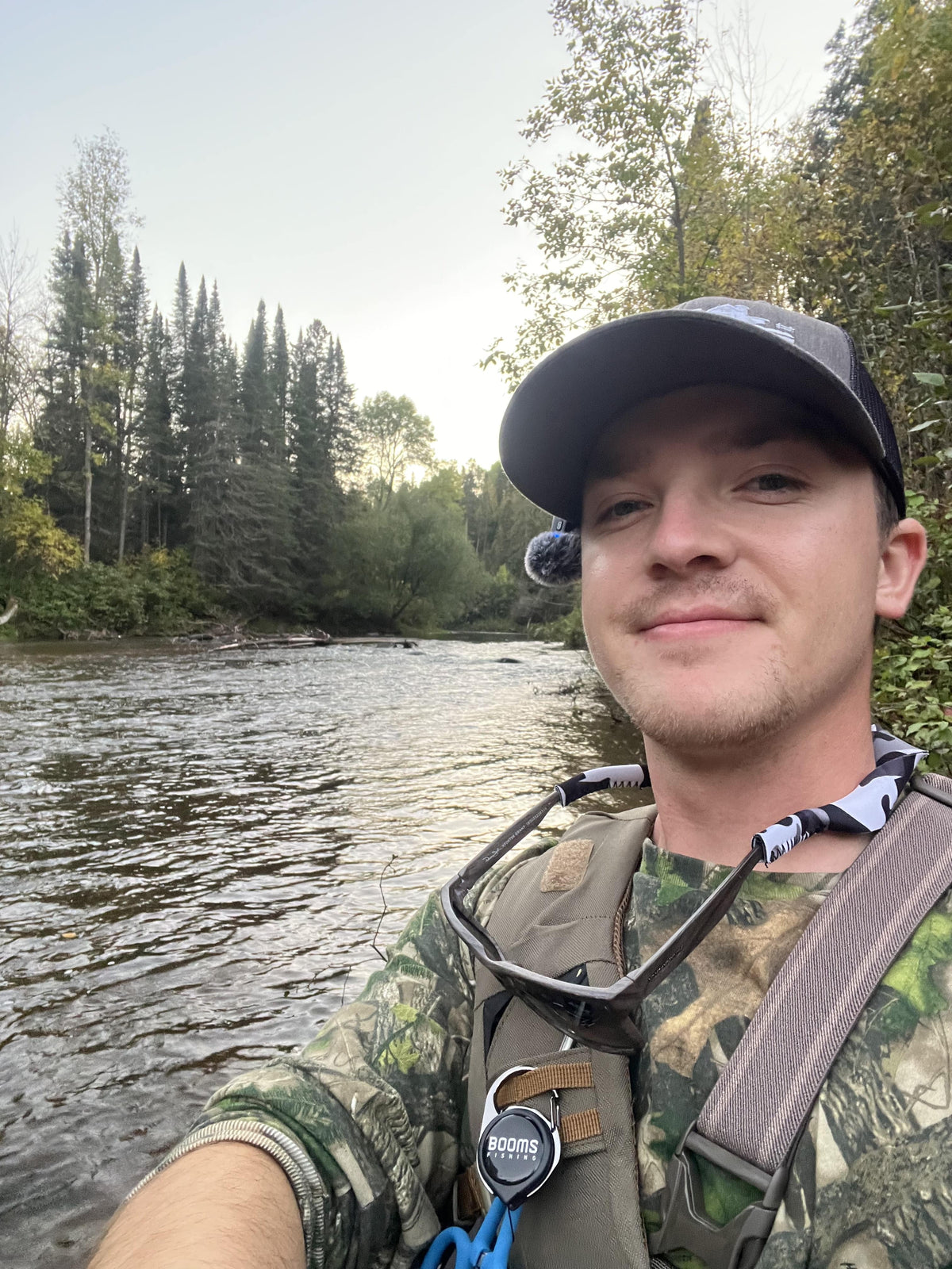 Man in camouflage clothing with a harness standing in the Brule river in northern Wisconsin with trees in the background.